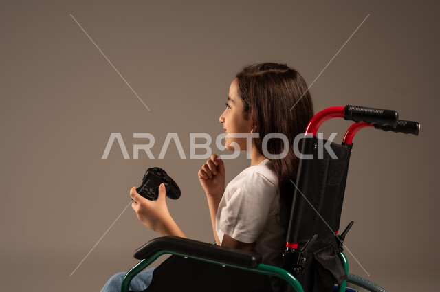 تجربة مثيرة في الأجهزة الحديثة الالكترونية، تحسين جودة حياة ذوي الاعاقة الحركية، نشاطات ترفيهية لذوي الهمم، التفاعل والاندماج مع ألعاب البلايستيشن, بورتريه لفتاة عربية خليجية سعودية ترتدي الزي الكاجوال تجلس على كرسي متحرك تحمل بيدها عصا التحكم،  خلفية رمادية