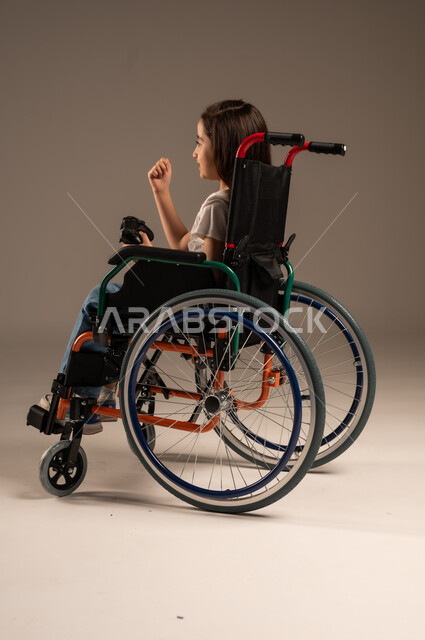 تجربة مثيرة في الأجهزة الحديثة الالكترونية، تحسين جودة حياة ذوي الاعاقة الحركية، نشاطات ترفيهية لذوي الهمم، التفاعل والاندماج مع ألعاب البلايستيشن, بورتريه لفتاة عربية خليجية سعودية ترتدي الزي الكاجوال تجلس على كرسي متحرك تحمل بيدها عصا التحكم،  خلفية رمادية