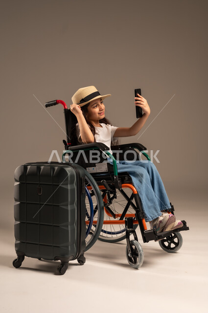 التقاط صورة سيلفي تذكارية, الاستعداد لقضاء إجازة صيفية ممتعة، التحدث مع الاقارب عبر الهاتف المحمول, بورتريه مقرب لفتاة عربية خليجية سعودية تجلس على كرسي متحرك تقوم بإجراء مكالمة فيديو وبجانبها حقيبة سفر, تأمين اجهزة ومعدات تساعد بالحركة والتنقل، خلفية رمادية
