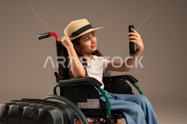 التقاط صورة سيلفي تذكارية, الاستعداد لقضاء إجازة صيفية ممتعة، التحدث مع الاقارب عبر الهاتف المحمول, بورتريه مقرب لفتاة عربية خليجية سعودية تجلس على كرسي متحرك تقوم بإجراء مكالمة فيديو وبجانبها حقيبة سفر, تأمين اجهزة ومعدات تساعد بالحركة والتنقل، خلفية رمادية
