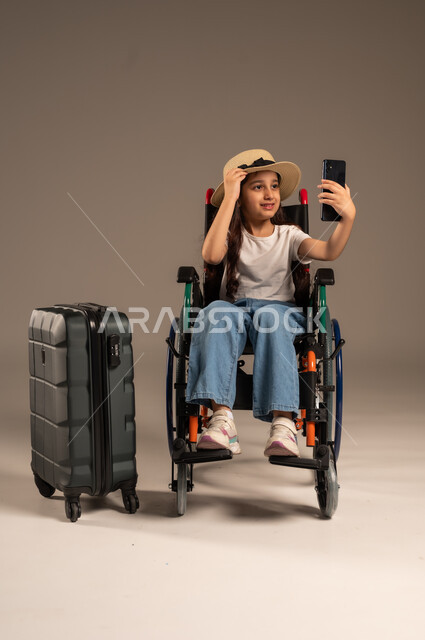 التقاط صورة سيلفي تذكارية, الاستعداد لقضاء إجازة صيفية ممتعة، التحدث مع الاقارب عبر الهاتف المحمول, بورتريه مقرب لفتاة عربية خليجية سعودية تجلس على كرسي متحرك تقوم بإجراء مكالمة فيديو وبجانبها حقيبة سفر, تأمين اجهزة ومعدات تساعد بالحركة والتنقل، خلفية رمادية