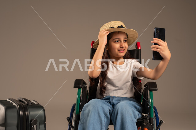 Taking a souvenir selfie, preparing for a fun summer vacation, talking to relatives on a mobile phone, close-up portrait of a Saudi Arabian Gulf girl sitting in a wheelchair making a video call with a suitcase next to her, securing devices and equipment that help with movement and mobility, gray background