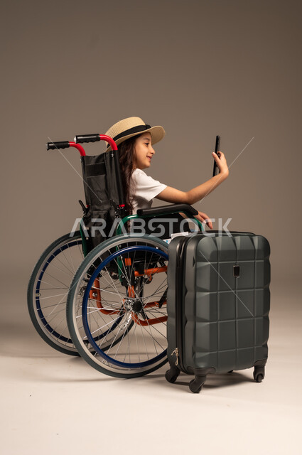 التقاط صورة سيلفي تذكارية, الاستعداد لقضاء إجازة صيفية ممتعة، التحدث مع الاقارب عبر الهاتف المحمول, بورتريه مقرب لفتاة عربية خليجية سعودية تجلس على كرسي متحرك تقوم بإجراء مكالمة فيديو وبجانبها حقيبة سفر, تأمين اجهزة ومعدات تساعد بالحركة والتنقل، خلفية رمادية