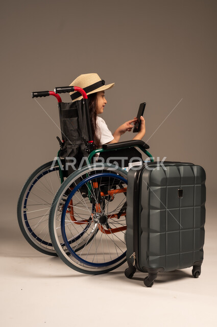 التقاط صورة سيلفي تذكارية, الاستعداد لقضاء إجازة صيفية ممتعة، التحدث مع الاقارب عبر الهاتف المحمول, بورتريه مقرب لفتاة عربية خليجية سعودية تجلس على كرسي متحرك تقوم بإجراء مكالمة فيديو وبجانبها حقيبة سفر, تأمين اجهزة ومعدات تساعد بالحركة والتنقل، خلفية رمادية