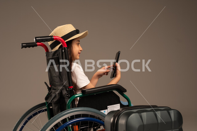التقاط صورة سيلفي تذكارية, الاستعداد لقضاء إجازة صيفية ممتعة، التحدث مع الاقارب عبر الهاتف المحمول, بورتريه مقرب لفتاة عربية خليجية سعودية تجلس على كرسي متحرك تقوم بإجراء مكالمة فيديو وبجانبها حقيبة سفر, تأمين اجهزة ومعدات تساعد بالحركة والتنقل، خلفية رمادية