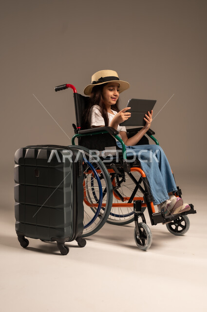 الاستعداد لقضاء إجازة صيفية ممتعة، التحدث مع الاقارب عبر الجهاز اللوحي, بورتريه مقرب لفتاة عربية خليجية سعودية تجلس على كرسي متحرك تقوم بإجراء مكالمة فيديو وبجانبها حقيبة سفر, تأمين اجهزة ومعدات تساعد بالحركة والتنقل، خلفية رمادية
