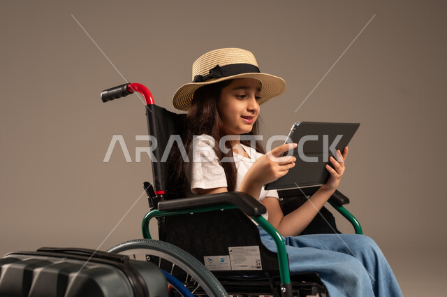الاستعداد لقضاء إجازة صيفية ممتعة، التحدث مع الاقارب عبر الجهاز اللوحي, بورتريه مقرب لفتاة عربية خليجية سعودية تجلس على كرسي متحرك تقوم بإجراء مكالمة فيديو وبجانبها حقيبة سفر, تأمين اجهزة ومعدات تساعد بالحركة والتنقل، خلفية رمادية