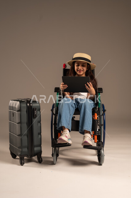 الاستعداد لقضاء إجازة صيفية ممتعة، التحدث مع الاقارب عبر الجهاز اللوحي, بورتريه مقرب لفتاة عربية خليجية سعودية تجلس على كرسي متحرك تقوم بإجراء مكالمة فيديو وبجانبها حقيبة سفر, تأمين اجهزة ومعدات تساعد بالحركة والتنقل، خلفية رمادية