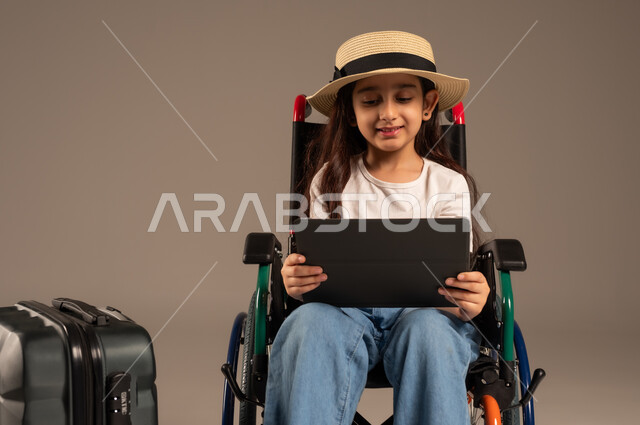 الاستعداد لقضاء إجازة صيفية ممتعة، التحدث مع الاقارب عبر الجهاز اللوحي, بورتريه مقرب لفتاة عربية خليجية سعودية تجلس على كرسي متحرك تقوم بإجراء مكالمة فيديو وبجانبها حقيبة سفر, تأمين اجهزة ومعدات تساعد بالحركة والتنقل، خلفية رمادية