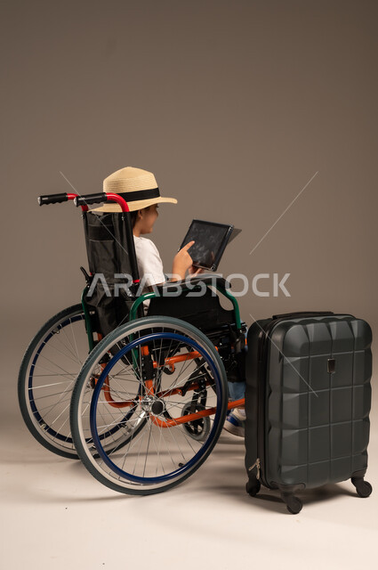 الاستعداد لقضاء إجازة صيفية ممتعة، التحدث مع الاقارب عبر الجهاز اللوحي, بورتريه مقرب لفتاة عربية خليجية سعودية تجلس على كرسي متحرك تقوم بإجراء مكالمة فيديو وبجانبها حقيبة سفر, تأمين اجهزة ومعدات تساعد بالحركة والتنقل، خلفية رمادية