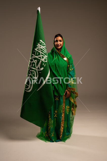 اليوم الوطني السعودي، المشاركة الأعياد الوطنية، بورتريه لامرأة عربية خليجية سعودية محجبة  ترتدي الشيلة والبشت وترفع علم السعودية بايماءات الفخر والاعتزاز بالوطن، يوم العلم السعودي 11 مارس، العيد الوطني 23 سبتمبر، الجسم بالطول الكامل، خلفية بيج