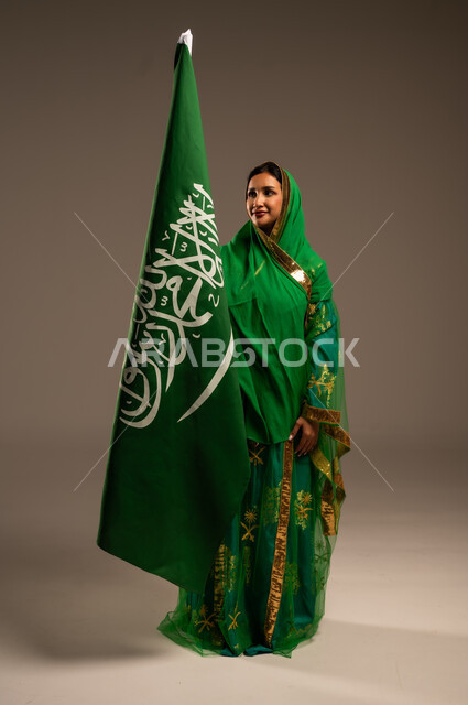 اليوم الوطني السعودي، المشاركة الأعياد الوطنية، بورتريه لامرأة عربية خليجية سعودية محجبة  ترتدي الشيلة والبشت وترفع علم السعودية بايماءات الفخر والاعتزاز بالوطن، يوم العلم السعودي 11 مارس، العيد الوطني 23 سبتمبر، الجسم بالطول الكامل، خلفية بيج