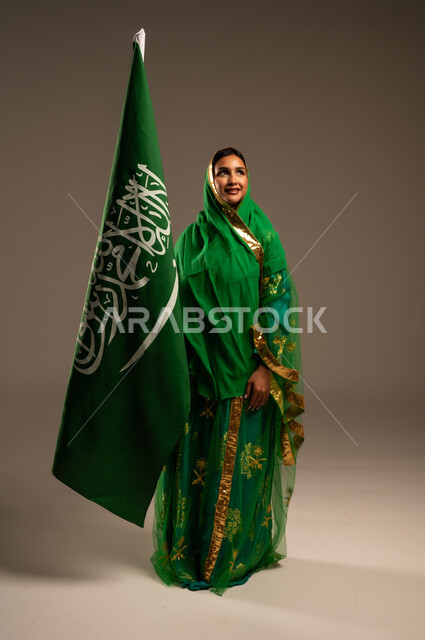 اليوم الوطني السعودي، المشاركة الأعياد الوطنية، بورتريه لامرأة عربية خليجية سعودية محجبة  ترتدي الشيلة والبشت وترفع علم السعودية بايماءات الفخر والاعتزاز بالوطن، يوم العلم السعودي 11 مارس، العيد الوطني 23 سبتمبر، الجسم بالطول الكامل، خلفية بيج