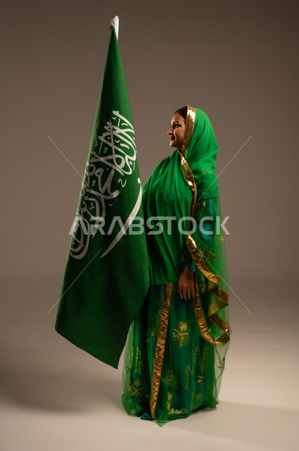 اليوم الوطني السعودي، المشاركة الأعياد الوطنية، بورتريه لامرأة عربية خليجية سعودية محجبة  ترتدي الشيلة والبشت وترفع علم السعودية بايماءات الفخر والاعتزاز بالوطن، يوم العلم السعودي 11 مارس، العيد الوطني 23 سبتمبر، الجسم بالطول الكامل، خلفية بيج