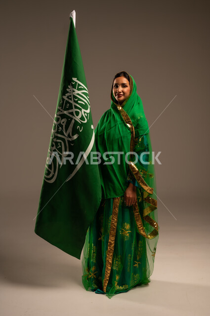 اليوم الوطني السعودي، المشاركة الأعياد الوطنية، بورتريه لامرأة عربية خليجية سعودية محجبة  ترتدي الشيلة والبشت وترفع علم السعودية بايماءات الفخر والاعتزاز بالوطن، يوم العلم السعودي 11 مارس، العيد الوطني 23 سبتمبر، الجسم بالطول الكامل، خلفية بيج