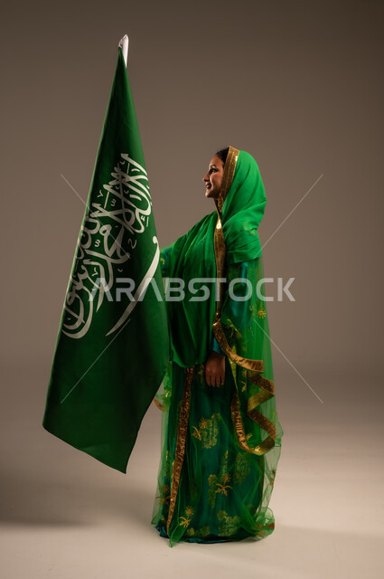 اليوم الوطني السعودي، المشاركة الأعياد الوطنية، بورتريه لامرأة عربية خليجية سعودية محجبة  ترتدي الشيلة والبشت وترفع علم السعودية بايماءات الفخر والاعتزاز بالوطن، يوم العلم السعودي 11 مارس، العيد الوطني 23 سبتمبر، الجسم بالطول الكامل، خلفية بيج