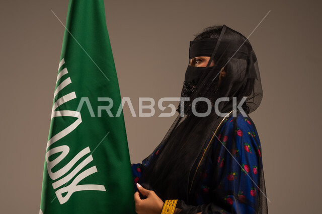 يوم بدينا 1727 م، ذكرى تأسيس الدولة السعودية الأولى 22 فبراير، اليوم الوطني السعودي، بورتريه لإمرأة عربية خليجية سعودية ترتدي زي المنطقة الوسطى وتمسك بيدها علم التأسيس, الاعتزاز بالبلد والانتماء اليه، خلفية بيج