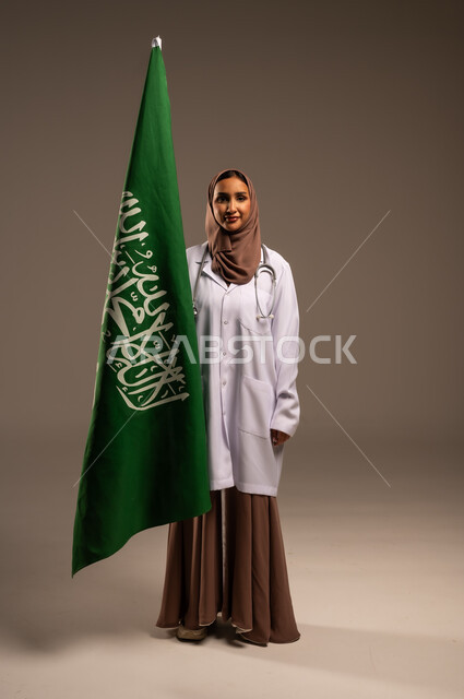  الاحتفال باليوم الوطني السعودي بفخر وعزة , بورتريه لطبيبة عربية خليجية سعودية ترتدي المعطف الطبي وسماعة الطبيب، ذكرى توحيد المملكة العربية السعودية , العلم السعودي راية التوحيد والاسلام , الجسم بالطول الكامل، خلفية بيج .