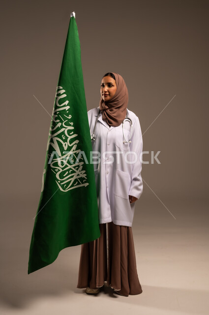 Celebrating Saudi National Day with pride and dignity, portrait of a Saudi Arabian Gulf female doctor wearing a medical coat and a stethoscope, anniversary of the unification of the Kingdom of Saudi Arabia, the Saudi flag, the banner of monotheism and Islam, full-length body, beige background.