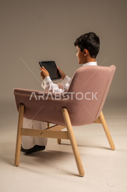 استخدام الأجهزة المحمولة في التعلم والترفيه، متابعة البرامج التعليمية والترفيهية ، بورتريه لولد عربي خليجي سعودي  يرتدي الثوب التقليدي يجلس على الكرسي ويقوم باستخدام  جهاز التابلت، خلفية بيج