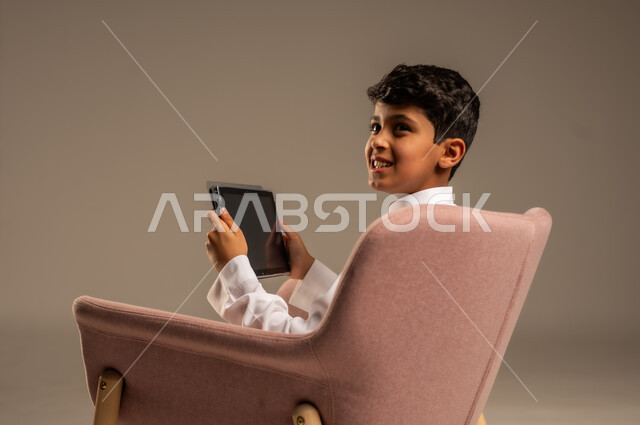 استخدام الأجهزة المحمولة في التعلم والترفيه، متابعة البرامج التعليمية والترفيهية ، بورتريه لولد عربي خليجي سعودي  يرتدي الثوب التقليدي يجلس على الكرسي ويقوم باستخدام  جهاز التابلت، خلفية بيج