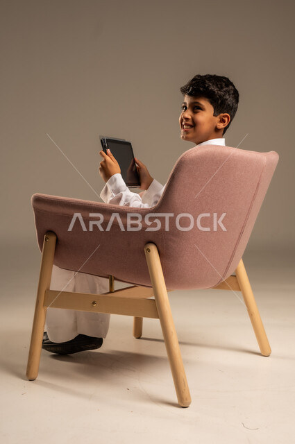 استخدام الأجهزة المحمولة في التعلم والترفيه، متابعة البرامج التعليمية والترفيهية ، بورتريه لولد عربي خليجي سعودي  يرتدي الثوب التقليدي يجلس على الكرسي ويقوم باستخدام  جهاز التابلت، خلفية بيج