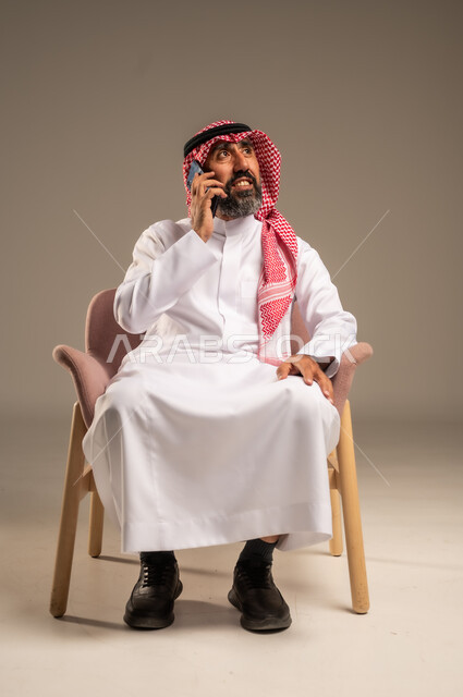 استخدام التطبيقات الحديثة واجراء المكالمات الهاتفية, بورتريه لرجل عربي سعودي خليجي كبير بالسن يرتدي الثوب التقليدي والشماغ يتحدث بالجوال, برامج التواصل الاجتماعي عبر الهاتف المحمول, خلفية بيج .