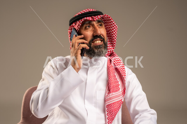استخدام التطبيقات الحديثة واجراء المكالمات الهاتفية, بورتريه لرجل عربي سعودي خليجي كبير بالسن يرتدي الثوب التقليدي والشماغ يتحدث بالجوال, برامج التواصل الاجتماعي عبر الهاتف المحمول, خلفية بيج .