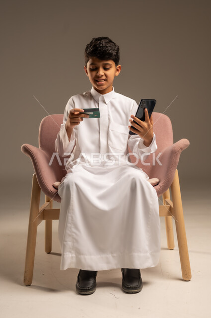 الشراء والدفع عن بعد عبر الاونلاين, استخدام بطاقة الإئتمان في التسوق الالكتروني، بورتريه مقرب لولد عربي سعودي خليجي يرتدي الثوب التقليدي يجلس على كرسي يمسك البطاقة البنكية ويقوم بتصفح تطبيقات المصارف والبنوك عبر الجوال، خلفية بيج