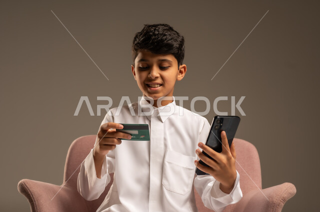 الشراء والدفع عن بعد عبر الاونلاين, استخدام بطاقة الإئتمان في التسوق الالكتروني، بورتريه مقرب لولد عربي سعودي خليجي يرتدي الثوب التقليدي يجلس على كرسي يمسك البطاقة البنكية ويقوم بتصفح تطبيقات المصارف والبنوك عبر الجوال، خلفية بيج