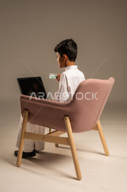 الشراء والدفع عن بعد عبر الاونلاين, استخدام بطاقة الإئتمان في التسوق الالكتروني، بورتريه من الخلف لولد عربي سعودي خليجي يرتدي الثوب التقليدي يجلس على كرسي يمسك البطاقة البنكية ويقوم بتصفح تطبيقات المصارف والبنوك عبر اللابتوب، خلفية بيج