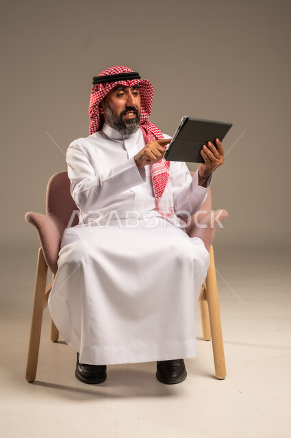 استخدام اجهزة تقنية حديثة، بورتريه لرجل عربي سعودي خليجي كبير بالسن يجلس على كرسي مريح يستخدم جهاز التابلت ، التسوق الإلكتروني الآمن، تصفح الإنترنت وإنجاز المهام عن بعد ، الشراء والبيع عبر الاونلاين، خلفية بيج