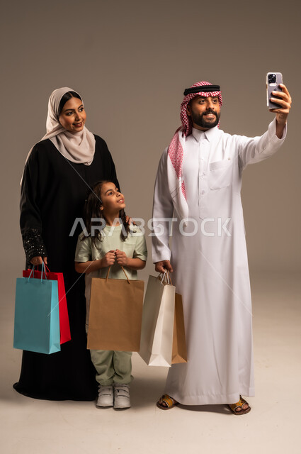 الاستمتاع بالتسوق برفقة الأسرة، التقاط صور السيلفي التذكارية باستخدام الهاتف المحمول، بورتريه لعائلة عربية خليجية سعودية بالملابس التقليدية يحملون الأكياس الورقية الملونة، أهمية قضاء الأوقات العائلية وتقوية روابط المحبة والمودة، الجسم بالطول الكامل، خلفية بيج