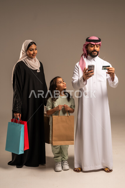 استخدام بطاقة الائتمان البنكية، الاستفادة من خدمات الدفع الالكتروني، الاستمتاع بالتسوق برفقة الأسرة وتقوية روابط المحبة والمودة، بورتريه مقرب لعائلة عربية خليجية سعودية بالملابس التقليدية يحملون الأكياس الورقية الملونة، الجسم بالطول الكامل، خلفية بيج