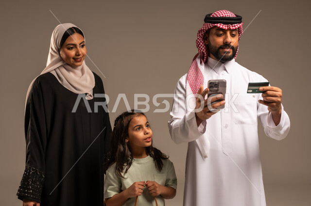 الاستفادة من خدمات الدفع الالكتروني، استخدام بطاقة الائتمان البنكية، تسهيل عمليات الشراء والحسابات، الاستمتاع بالتسوق برفقة الأسرة وتقوية روابط المحبة والمودة، بورتريه مقرب لعائلة عربية خليجية سعودية بالملابس التقليدية تقضي وقت عائلي ممتع، خلفية بيج