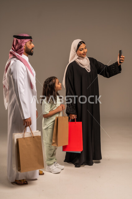 Enjoying shopping with family, taking souvenir selfies using mobile phone, portrait of a Saudi Arabian Gulf family in traditional clothes holding colorful paper bags, importance of spending family time and strengthening bonds of love and affection, full body, beige background