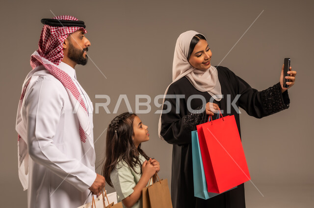 التقاط صور السيلفي التذكارية باستخدام الهاتف المحمول، الاستمتاع بالتسوق برفقة الأسرة، بورتريه مقرب لعائلة عربية خليجية سعودية بالملابس التقليدية يحملون الأكياس الورقية الملونة، أهمية قضاء الأوقات العائلية وتقوية روابط المحبة والمودة، خلفية بيج