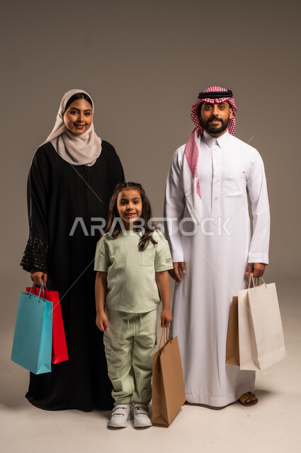  الوقوف والنظر إلى الكاميرا بإيماءات السعادة والسرور، الاستمتاع بالتسوق برفقة الأسرة وتقوية روابط المحبة والمودة، بورتريه لعائلة عربية خليجية سعودية بالملابس التقليدية يحملون الأكياس الورقية الملونة، شراء الهدايا والمستلزمات، الجسم بالطول الكامل، خلفية بيج