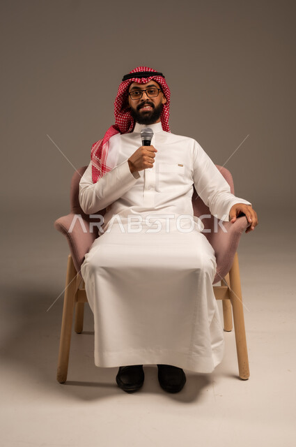 التواصل الفعال وتبادل الحوارات والنقاشات، إجراء المقابلات وطرح الأسئلة، بورتريه لرجل عربي خليجي سعودي يرتدي الثوب التقليدي والشماغ يجلس على الكرسي يحمل بيده مكبر الصوت يقوم بإلقاء الخطابات وينظر إلى الكاميرا، خلفية رمادية