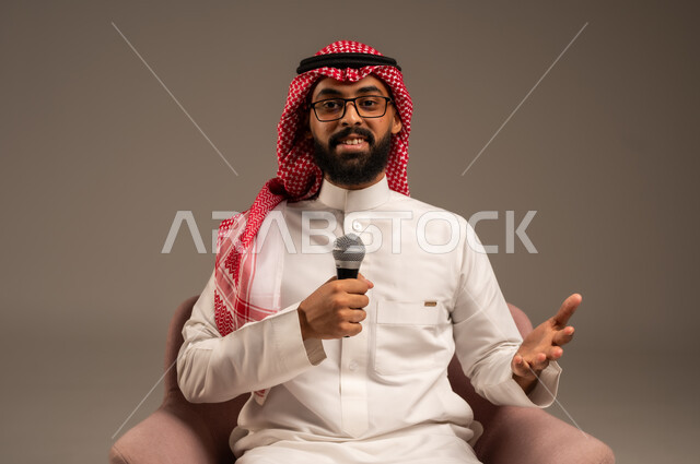 التواصل الفعال وتبادل الحوارات والنقاشات، إجراء المقابلات وطرح الأسئلة، بورتريه مقرب لرجل عربي خليجي سعودي يرتدي الثوب التقليدي والشماغ يجلس على الكرسي يحمل بيده مكبر الصوت يقوم بإلقاء الخطابات وينظر إلى الكاميرا، خلفية رمادية