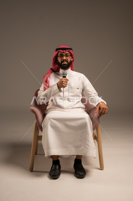 التواصل الفعال وتبادل الحوارات والنقاشات، إجراء المقابلات وطرح الأسئلة، بورتريه لرجل عربي خليجي سعودي يرتدي الثوب التقليدي والشماغ يجلس على الكرسي يحمل بيده مكبر الصوت يقوم بإلقاء الخطابات وينظر إلى الكاميرا، خلفية رمادية