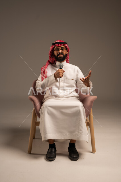التواصل الفعال وتبادل الحوارات والنقاشات، إجراء المقابلات وطرح الأسئلة، بورتريه لرجل عربي خليجي سعودي يرتدي الثوب التقليدي والشماغ يجلس على الكرسي يحمل بيده مكبر الصوت يقوم بإلقاء الخطابات وينظر إلى الكاميرا، خلفية رمادية