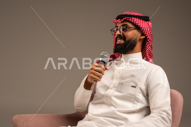 Interviewing and asking questions, close-up portrait of a Saudi Arabian Gulf man wearing traditional thobe and shemagh sitting on a chair holding a loudspeaker and giving speeches, effective communication and exchanging dialogues and discussions, gray background