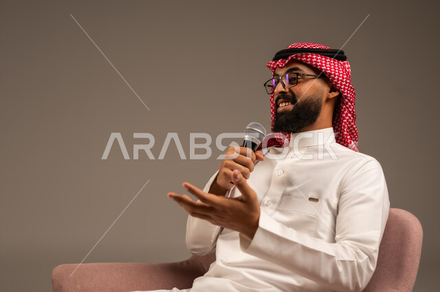 إجراء المقابلات وطرح الأسئلة، بورتريه مقرب لرجل عربي خليجي سعودي يرتدي الثوب التقليدي والشماغ يجلس على الكرسي يحمل بيده مكبر الصوت ويقوم بإلقاء الخطابات، التواصل الفعال وتبادل الحوارات والنقاشات، خلفية رمادية
