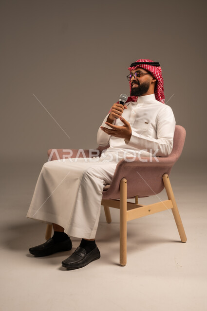 إجراء المقابلات وطرح الأسئلة، بورتريه لرجل عربي خليجي سعودي يرتدي الثوب التقليدي والشماغ يجلس على الكرسي يحمل بيده مكبر الصوت ويقوم بإلقاء الخطابات، التواصل الفعال وتبادل الحوارات والنقاشات، خلفية رمادية