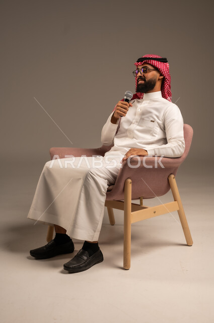 إجراء المقابلات وطرح الأسئلة، بورتريه لرجل عربي خليجي سعودي يرتدي الثوب التقليدي والشماغ يجلس على الكرسي يحمل بيده مكبر الصوت ويقوم بإلقاء الخطابات، التواصل الفعال وتبادل الحوارات والنقاشات، خلفية رمادية