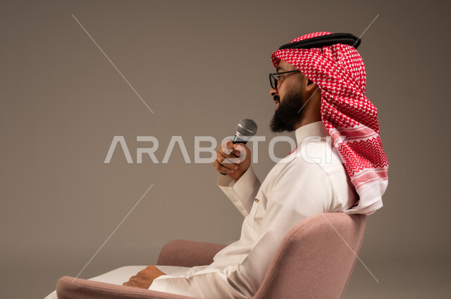 إجراء المقابلات وطرح الأسئلة، بورتريه مقرب من الجانب لرجل عربي خليجي سعودي يرتدي الثوب التقليدي والشماغ يجلس على الكرسي يحمل بيده مكبر الصوت ويقوم بإلقاء الخطابات، التواصل الفعال وتبادل الحوارات والنقاشات، خلفية رمادية