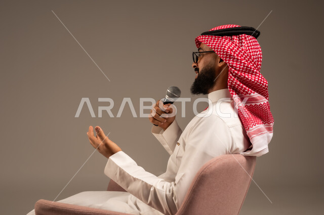 إجراء المقابلات وطرح الأسئلة، بورتريه مقرب من الجانب لرجل عربي خليجي سعودي يرتدي الثوب التقليدي والشماغ يجلس على الكرسي يحمل بيده مكبر الصوت ويقوم بإلقاء الخطابات، التواصل الفعال وتبادل الحوارات والنقاشات، خلفية رمادية