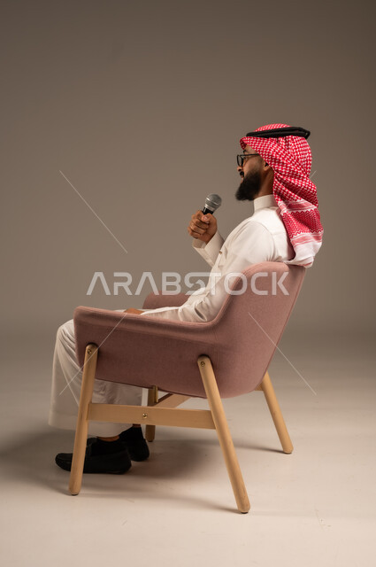 إجراء المقابلات وطرح الأسئلة، بورتريه من الجانب لرجل عربي خليجي سعودي يرتدي الثوب التقليدي والشماغ يجلس على الكرسي يحمل بيده مكبر الصوت ويقوم بإلقاء الخطابات، التواصل الفعال وتبادل الحوارات والنقاشات، خلفية رمادية