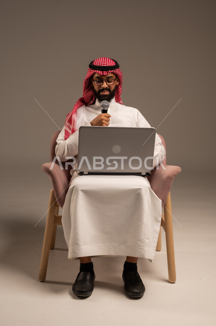 إجراء المقابلات وطرح الأسئلة، استخدام الأجهزة التقنية الحديثة، بورتريه لرجل عربي خليجي سعودي يرتدي الثوب التقليدي والشماغ يجلس على الكرسي يستخدم الحاسوب المحمول ويحمل بيده مكبر الصوت يقوم بإلقاء الخطابات، خلفية رمادية